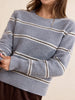 A close-up of a woman wearing a chunky knit crewneck sweater with horizontal stripes in light blue, cream, and dark brown, paired with a gold necklace and brown bottoms.