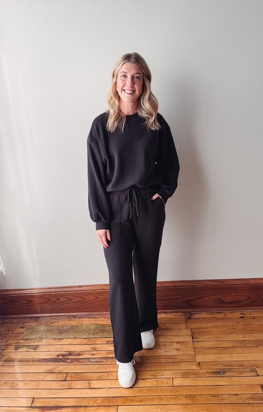 Woman wearing a black outfit standing on a wooden floor against a white wall