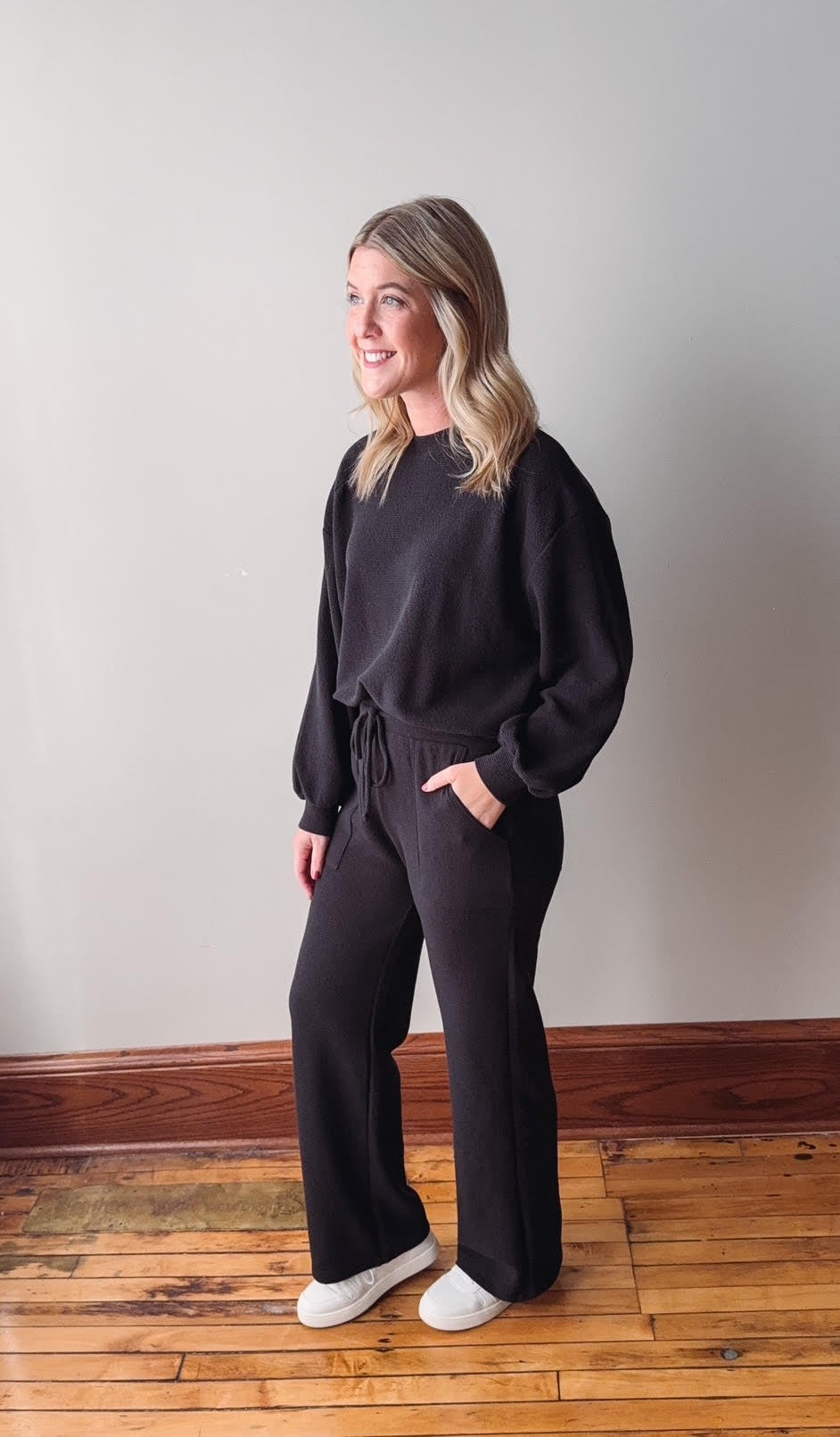 Woman wearing a black outfit standing on a wooden floor with a plain wall background