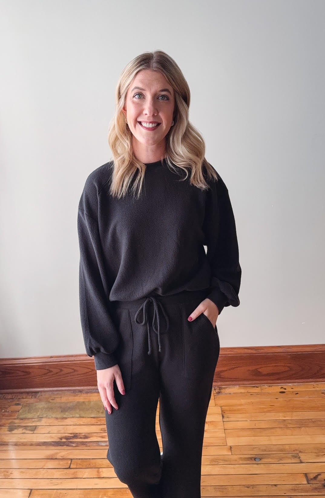 Woman wearing a black outfit standing in a room with wooden flooring and a white wall.