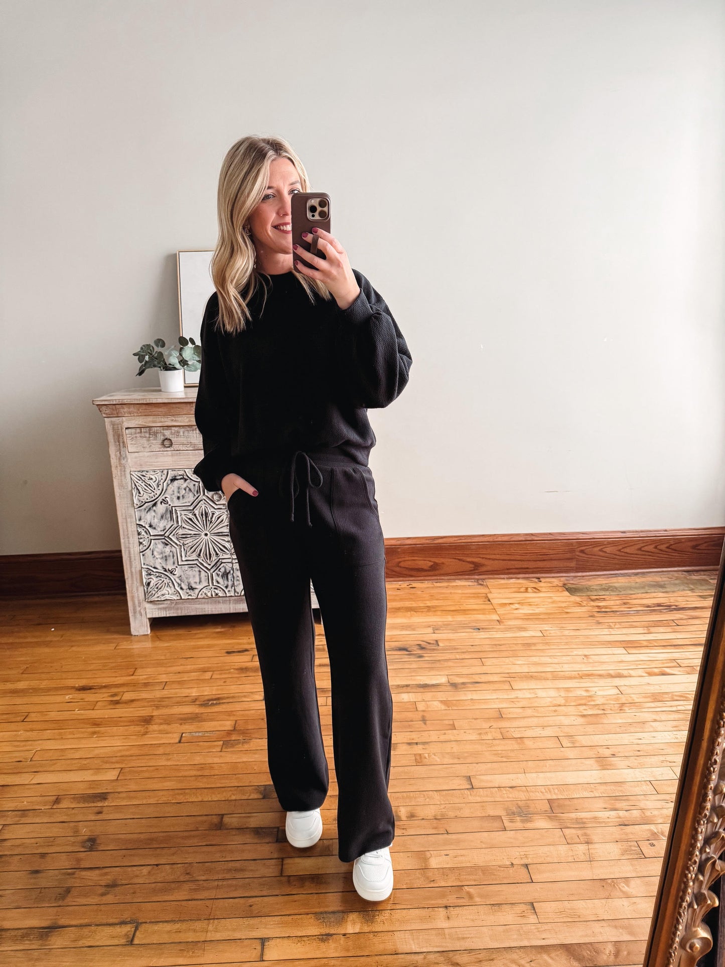 Person wearing a black outfit taking a mirror selfie in a room with wooden flooring and a decorative cabinet.