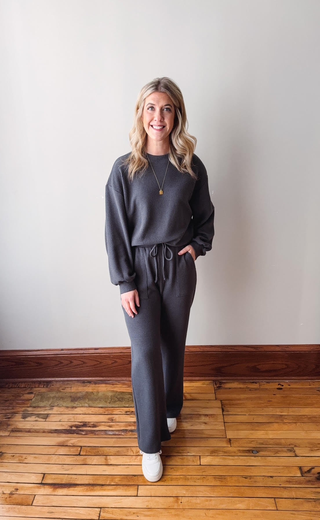 Woman wearing a gray outfit standing on a wooden floor with a white wall background