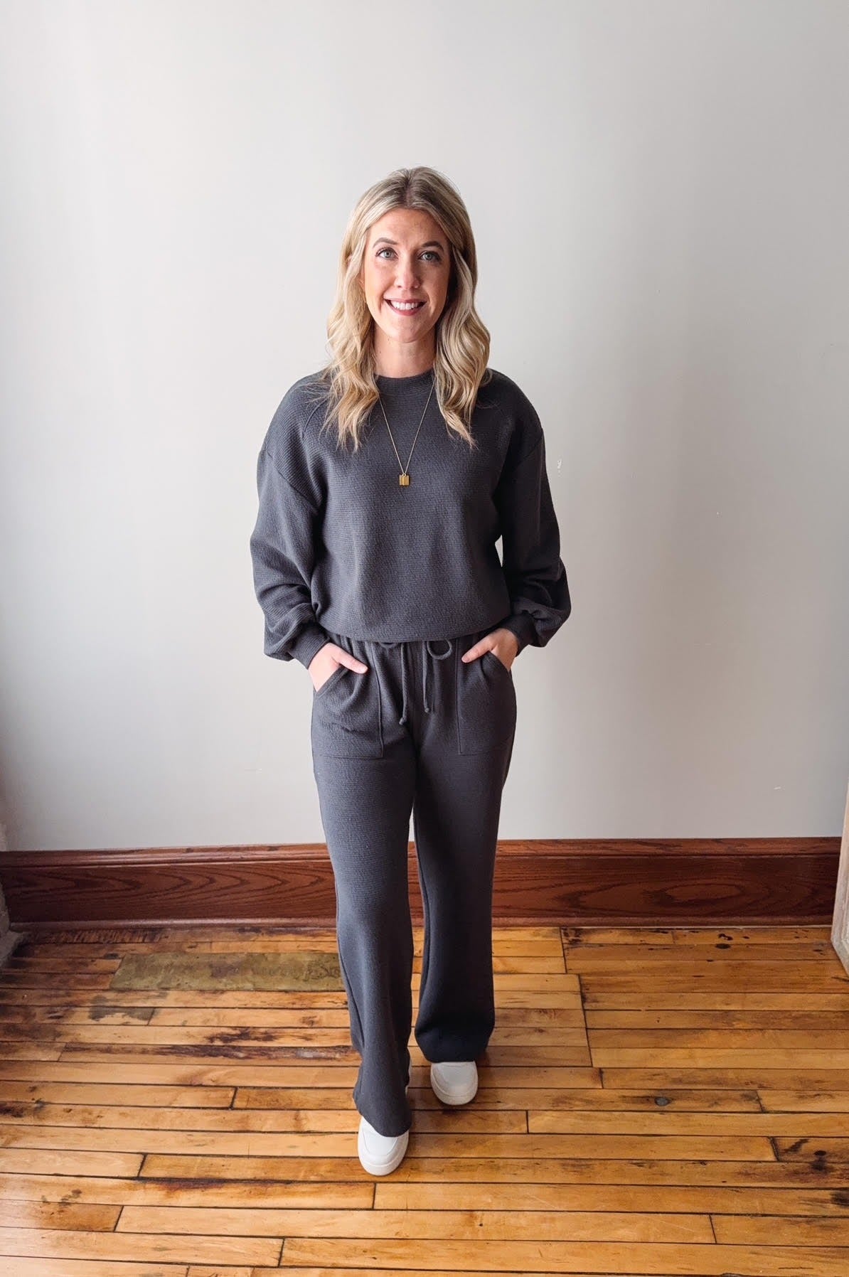 Woman wearing a gray outfit standing on a wooden floor with a white wall background