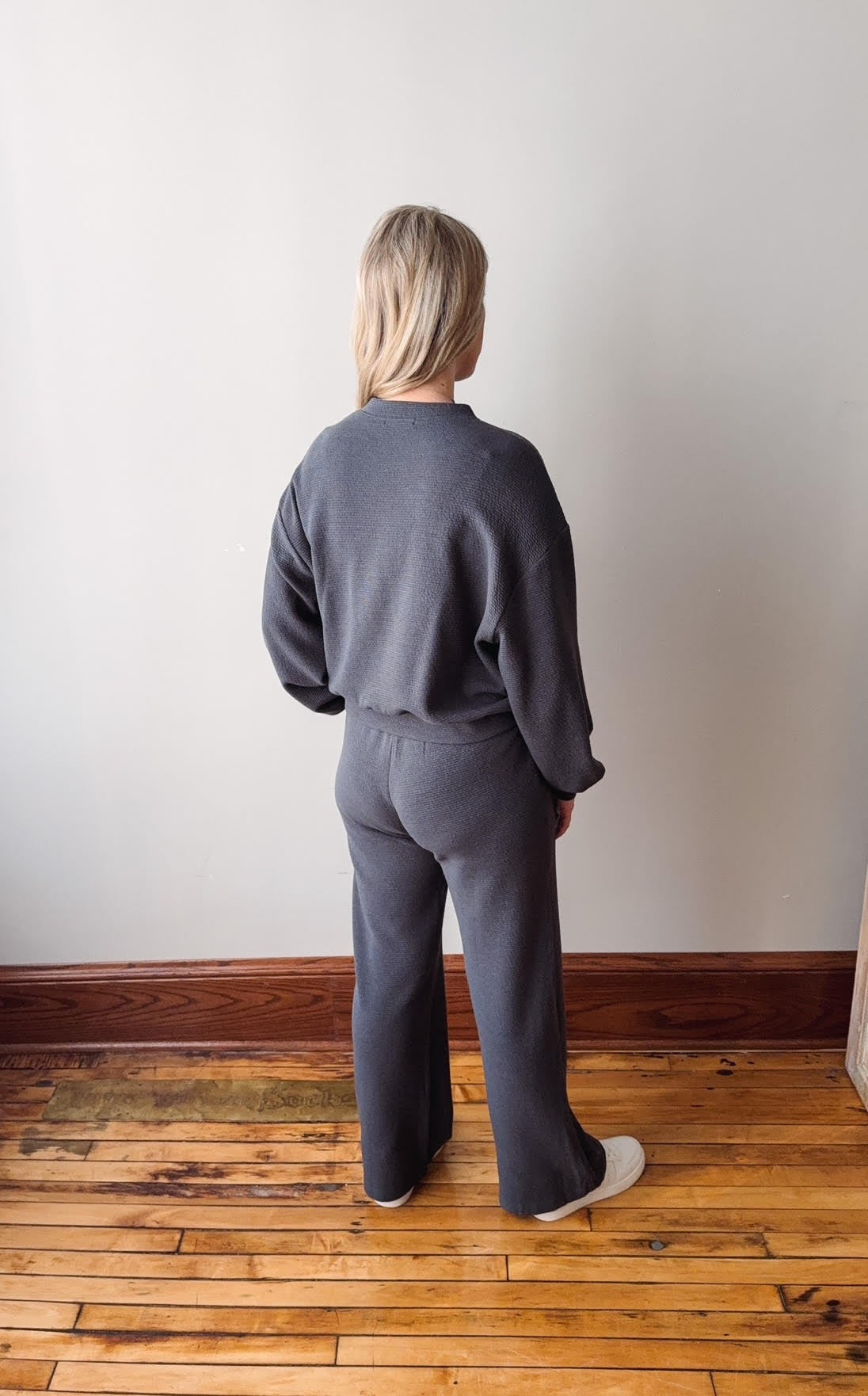 Person wearing a gray outfit standing on a wooden floor against a white wall