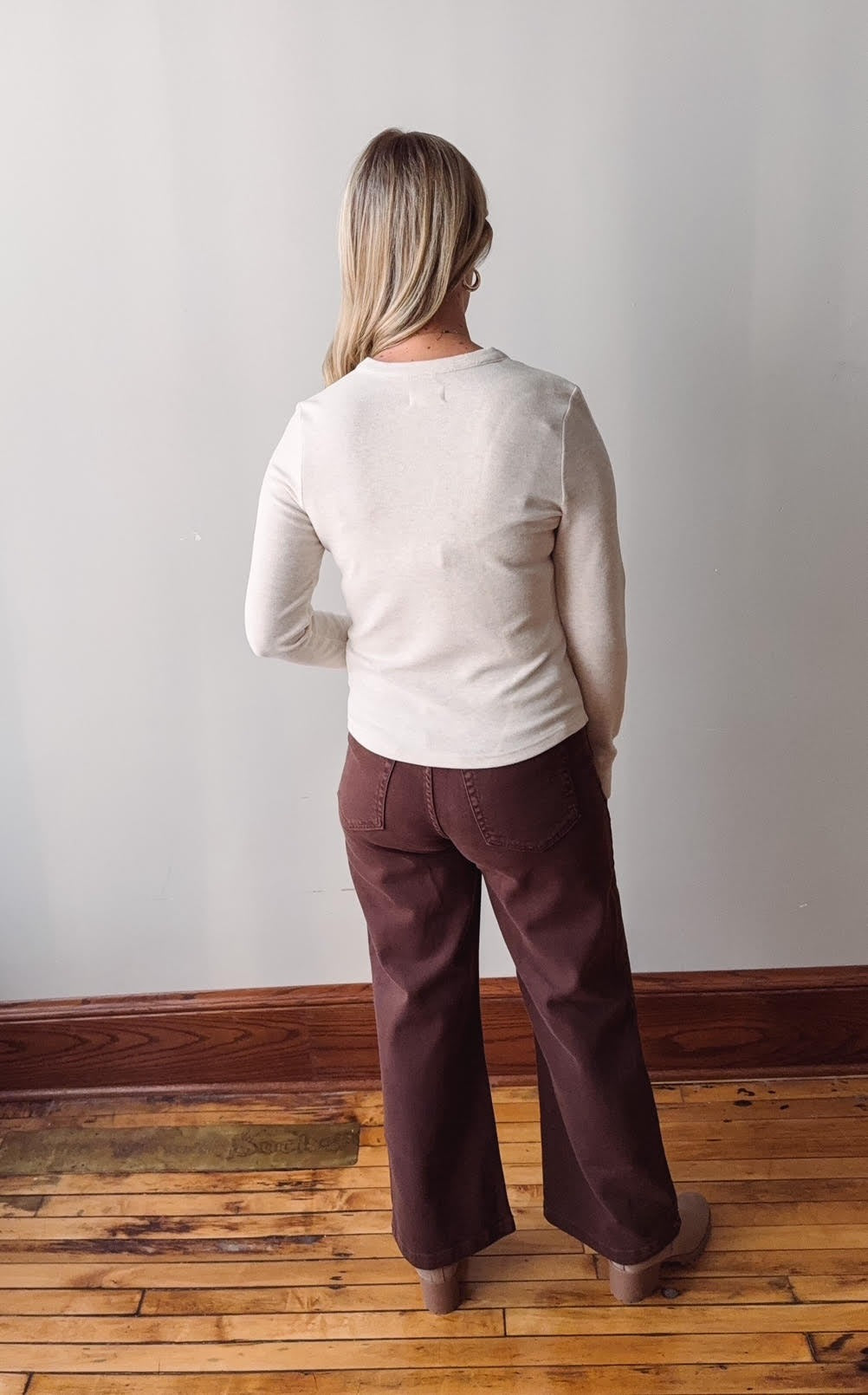 Person wearing a beige sweater and brown pants standing against a plain wall.