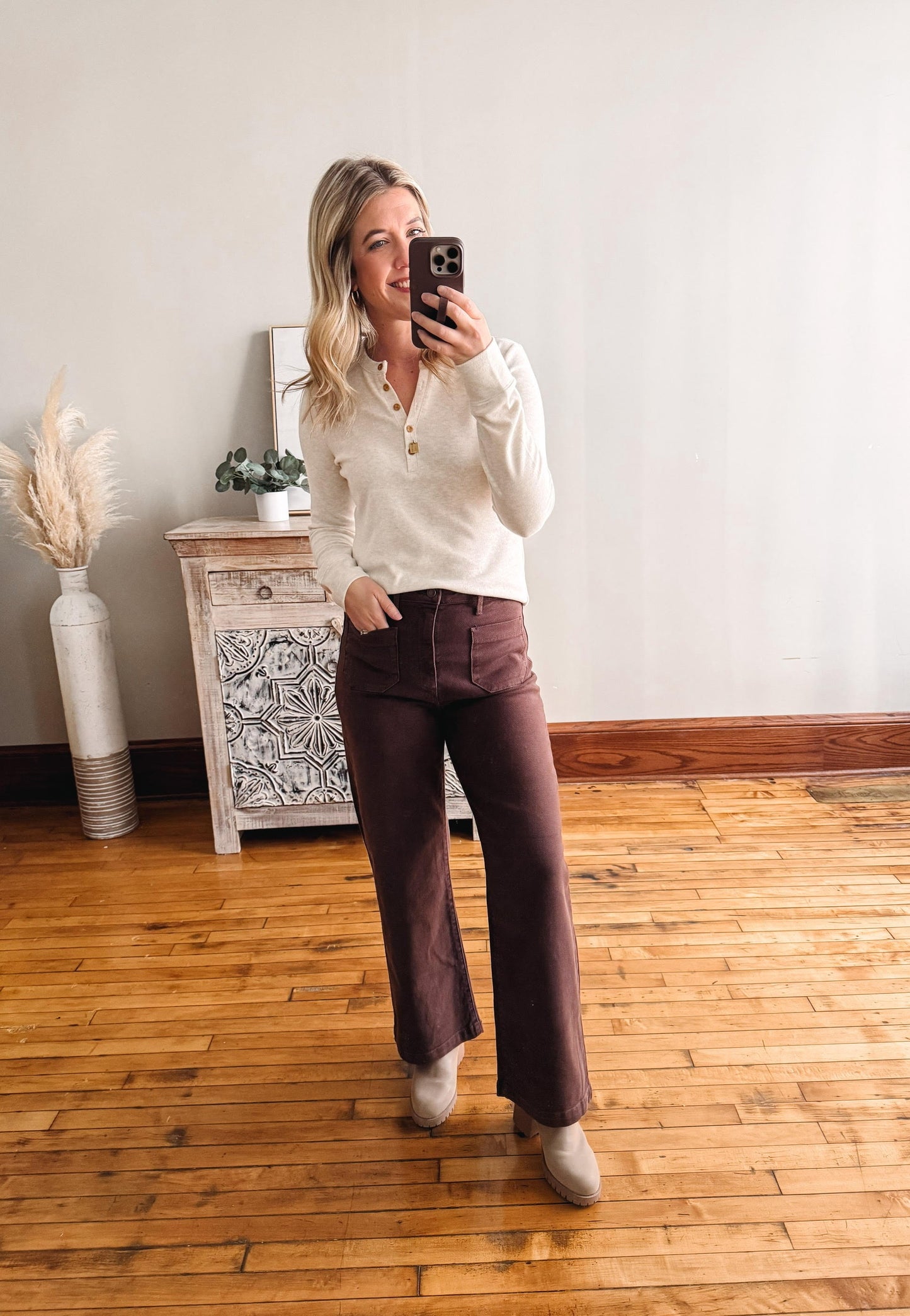 Woman taking a mirror selfie wearing a white blouse and brown pants in a room with wooden flooring.