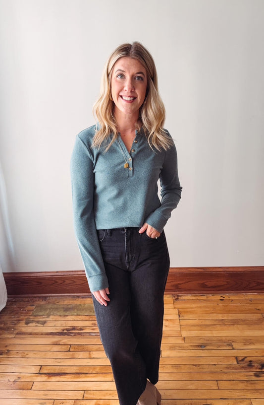 Woman wearing a light blue sweater and dark pants standing in a room with wooden flooring and a white wall.