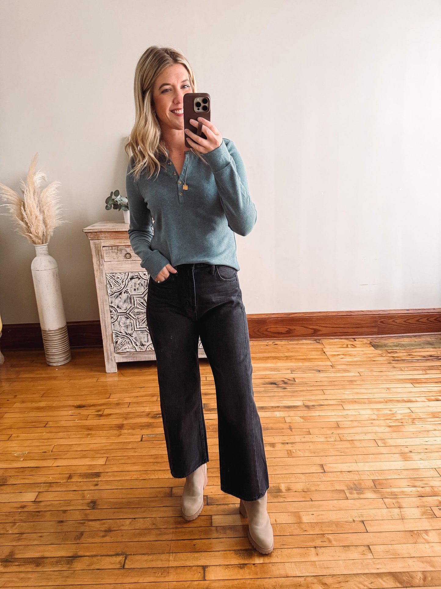 Woman taking a mirror selfie wearing a blue sweater and black pants in a room with wooden flooring.