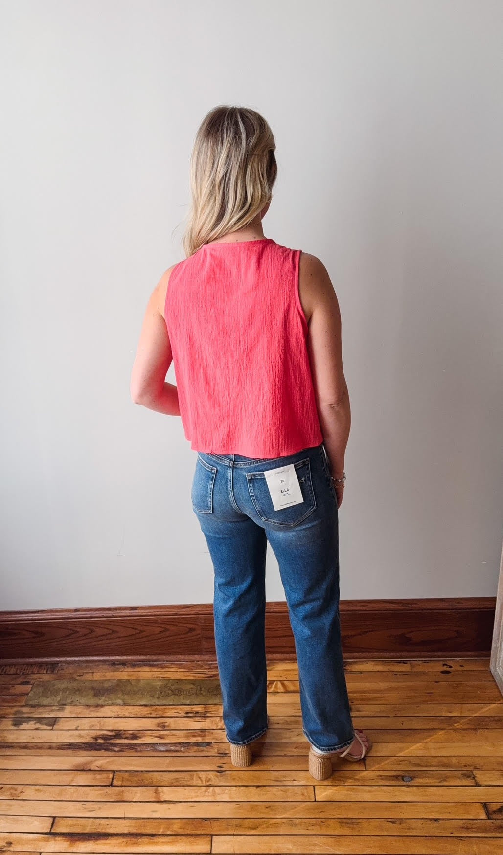 Person wearing a red sleeveless top and blue jeans standing on a wooden floor with a white wall background.