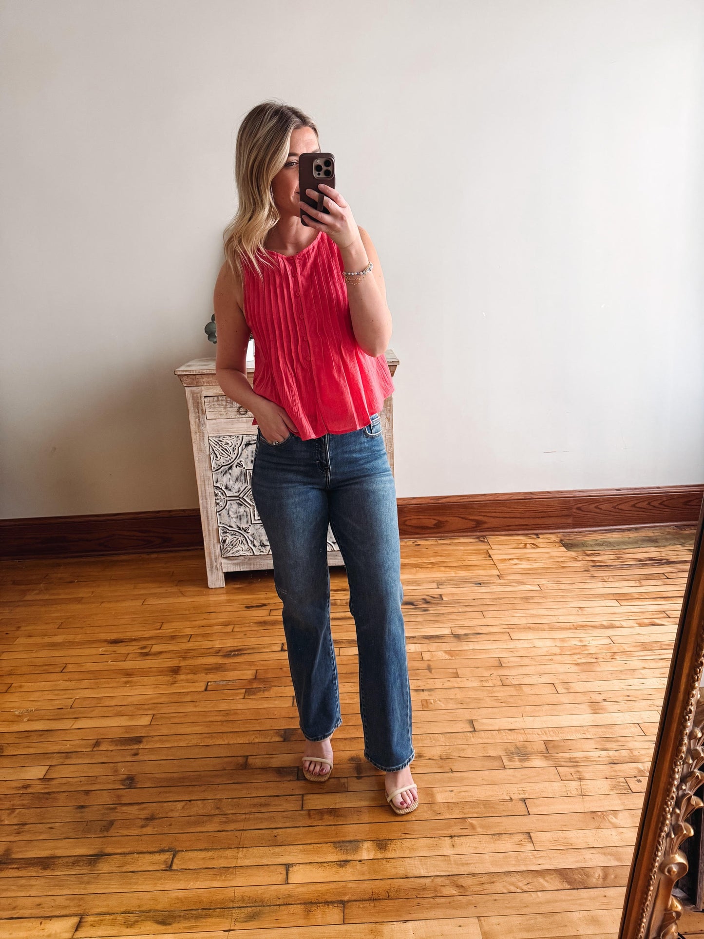 Woman taking a mirror selfie wearing a pink top and blue jeans in a room with wooden flooring.