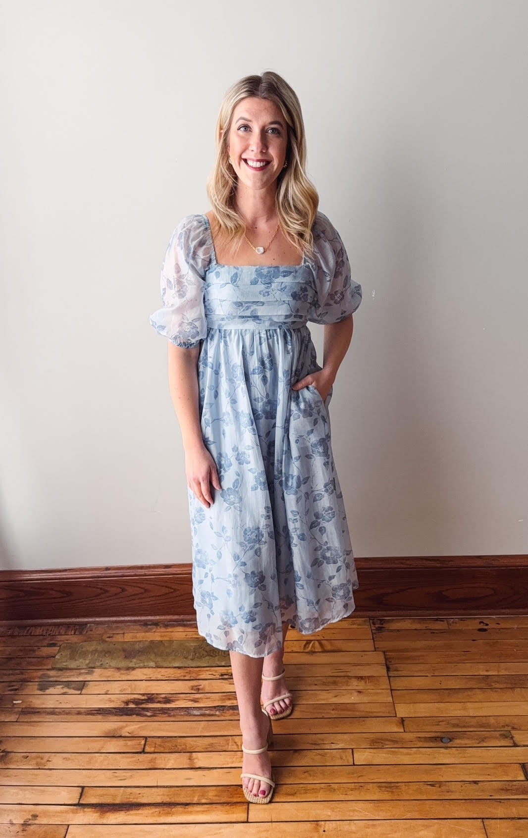 Woman wearing a floral dress standing on a wooden floor with a white wall background