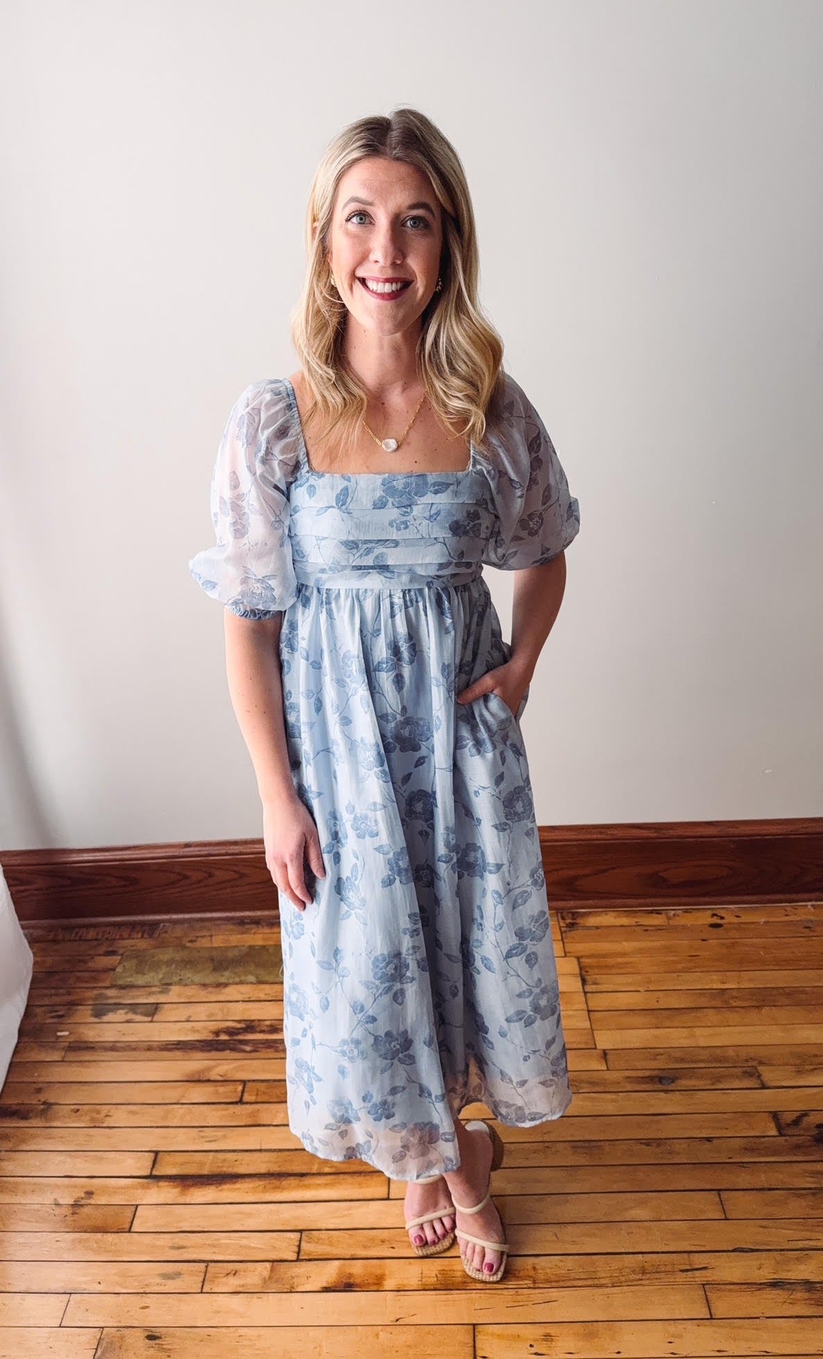 Woman wearing a blue floral dress standing on a wooden floor with a white wall background