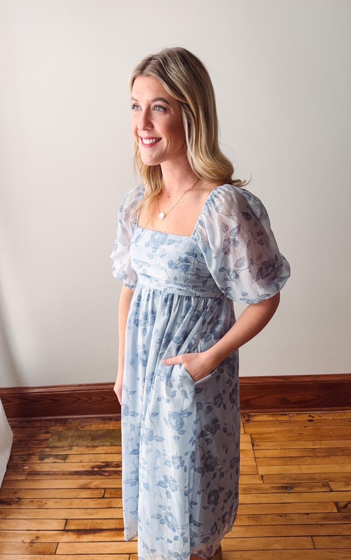 Woman wearing a light blue floral dress standing in a room with wooden flooring and a white wall.