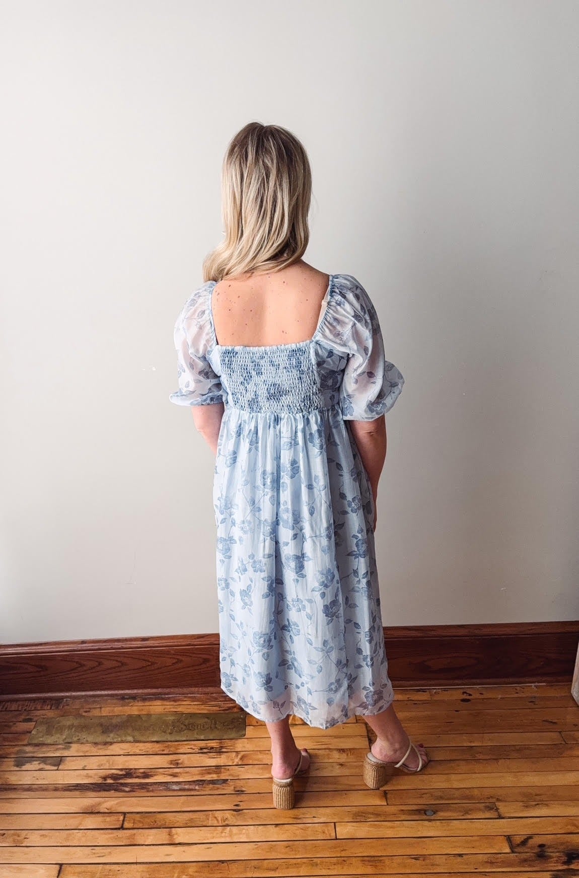 Woman wearing a blue floral dress standing on a wooden floor against a white wall.
