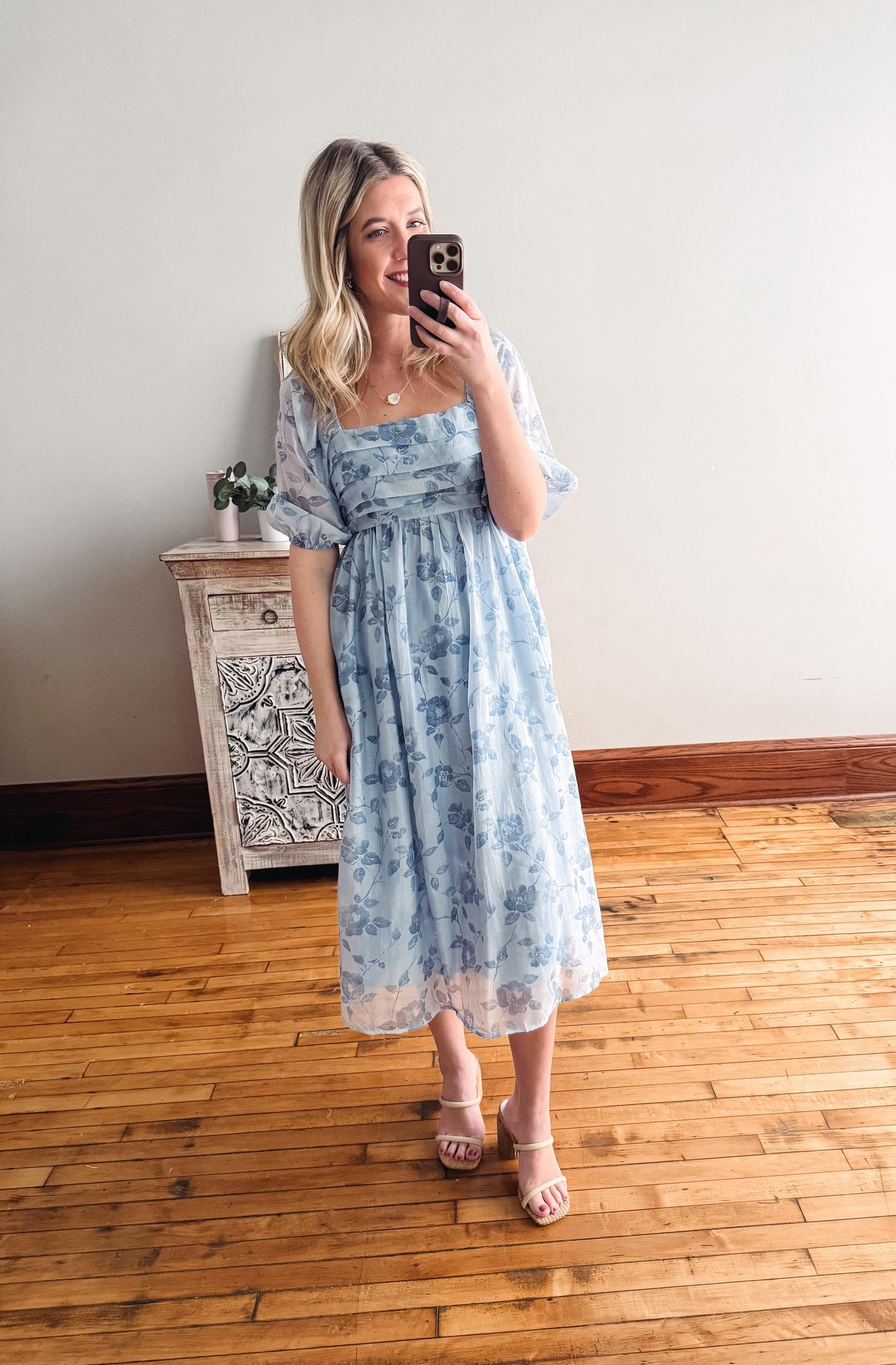 Woman taking a mirror selfie wearing a light blue floral dress in a room with wooden flooring and a white wall.