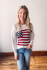 A woman smiling and wearing a white sweater with a bold American flag design in the center. She is also wearing blue jeans.