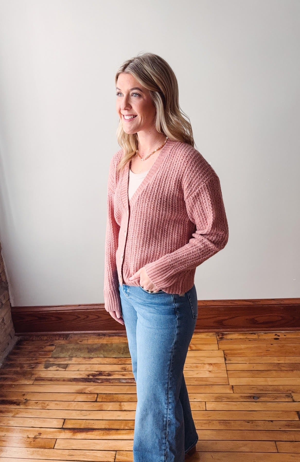 Woman wearing a pink cardigan and blue jeans standing in a room with wooden flooring and white walls.