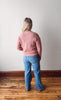 Person wearing a pink sweater and blue jeans standing in a room with wooden flooring and a white wall.