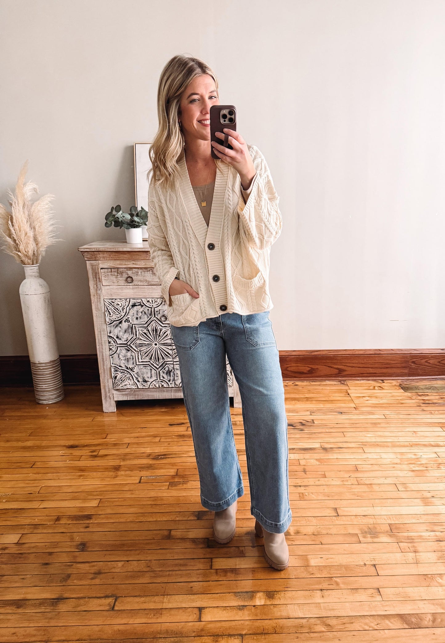 Woman taking a mirror selfie wearing a cream cardigan and blue jeans in a room with wooden flooring.