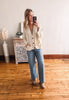 Woman taking a mirror selfie wearing a cream cardigan and blue jeans in a room with wooden flooring.