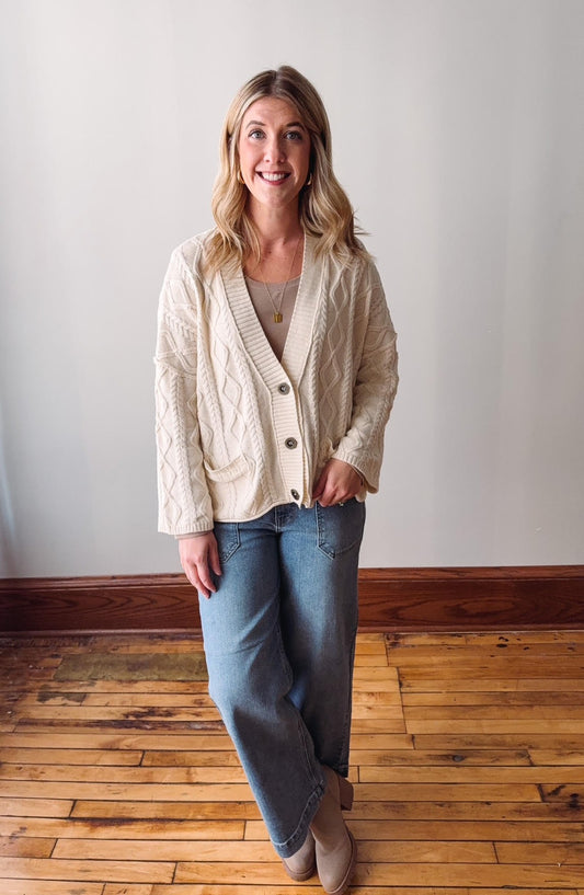 Woman wearing a cream cardigan and blue jeans standing on a wooden floor.