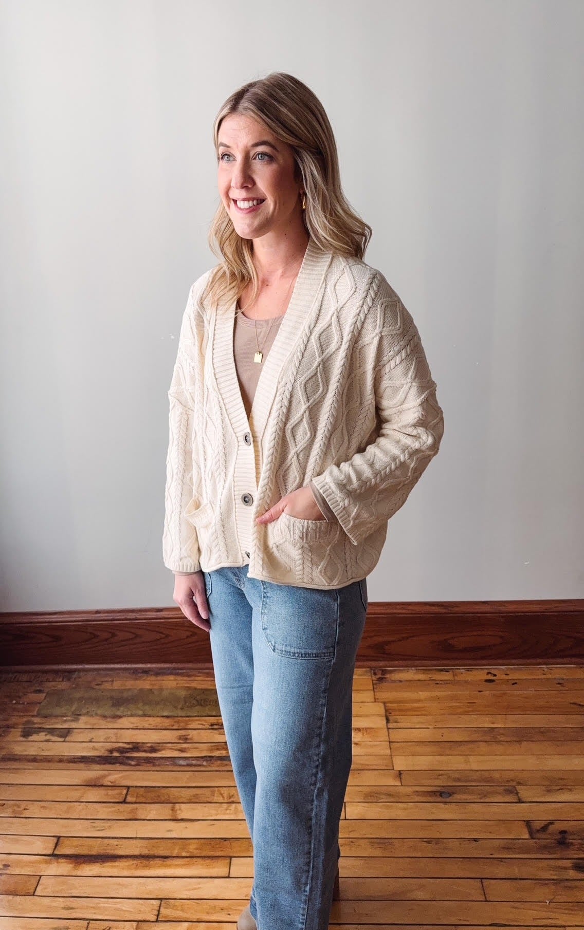 Woman wearing a cream cardigan and blue jeans standing on a wooden floor.