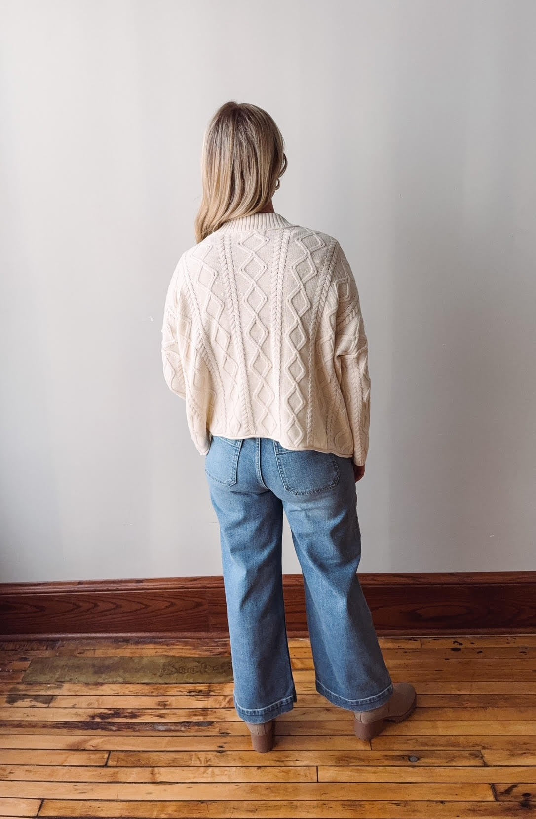 Person wearing a cream sweater and blue jeans standing against a plain white wall on a wooden floor.