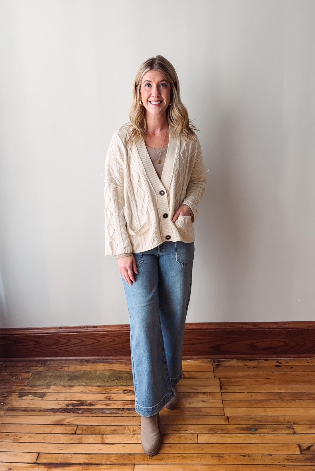 Woman wearing a beige cardigan, light blue jeans, and gray shoes standing on a wooden floor against a white wall.