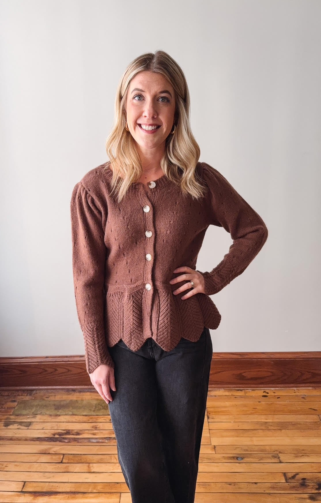 Woman wearing a brown knitted cardigan standing on a wooden floor with a white wall background