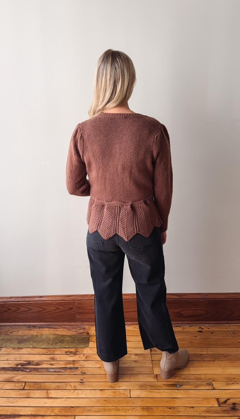 Person wearing a brown knit sweater with a scalloped hem and dark pants on a wooden floor.