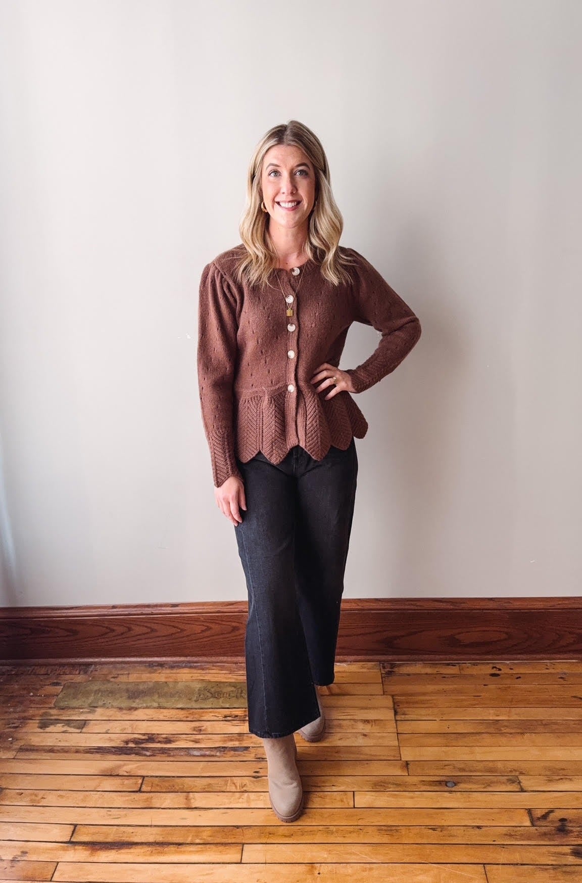 Woman wearing a brown cardigan and dark pants standing on a wooden floor against a white wall.