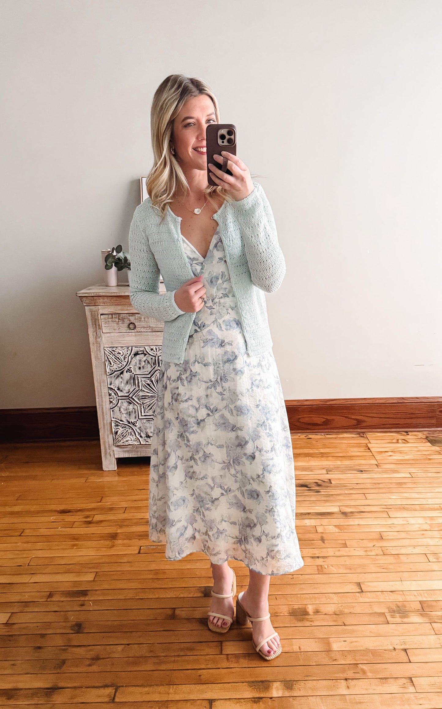 Woman taking a mirror selfie wearing a floral dress and gray cardigan in a room with wooden flooring.