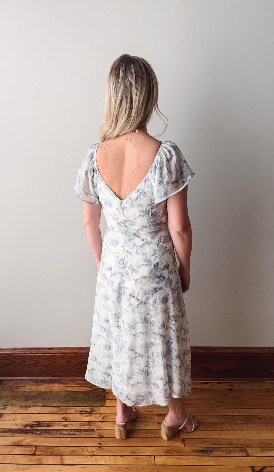 Woman wearing a floral dress standing on a wooden floor with a plain wall background