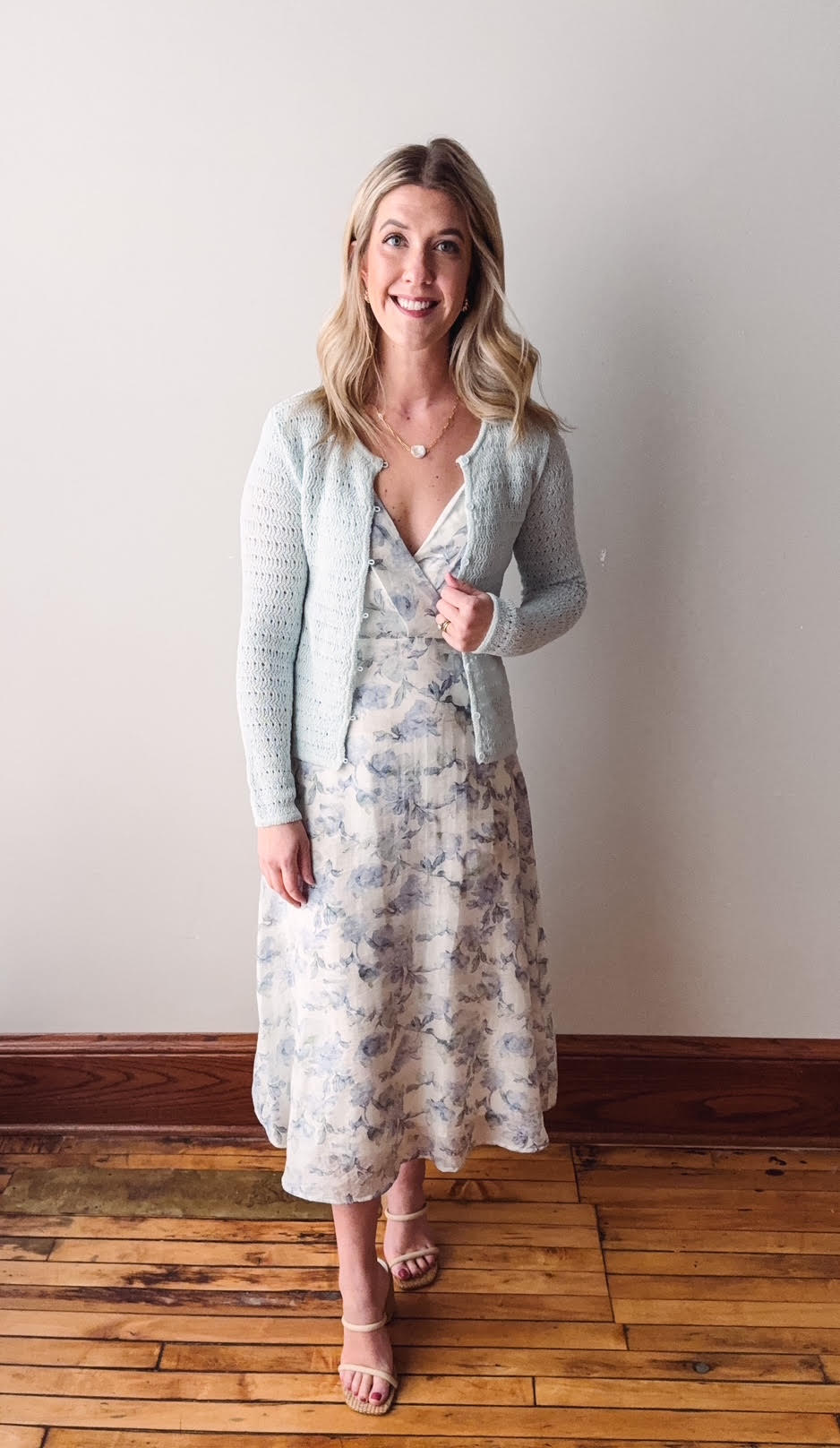 Woman wearing a floral dress and gray cardigan standing against a plain wall.