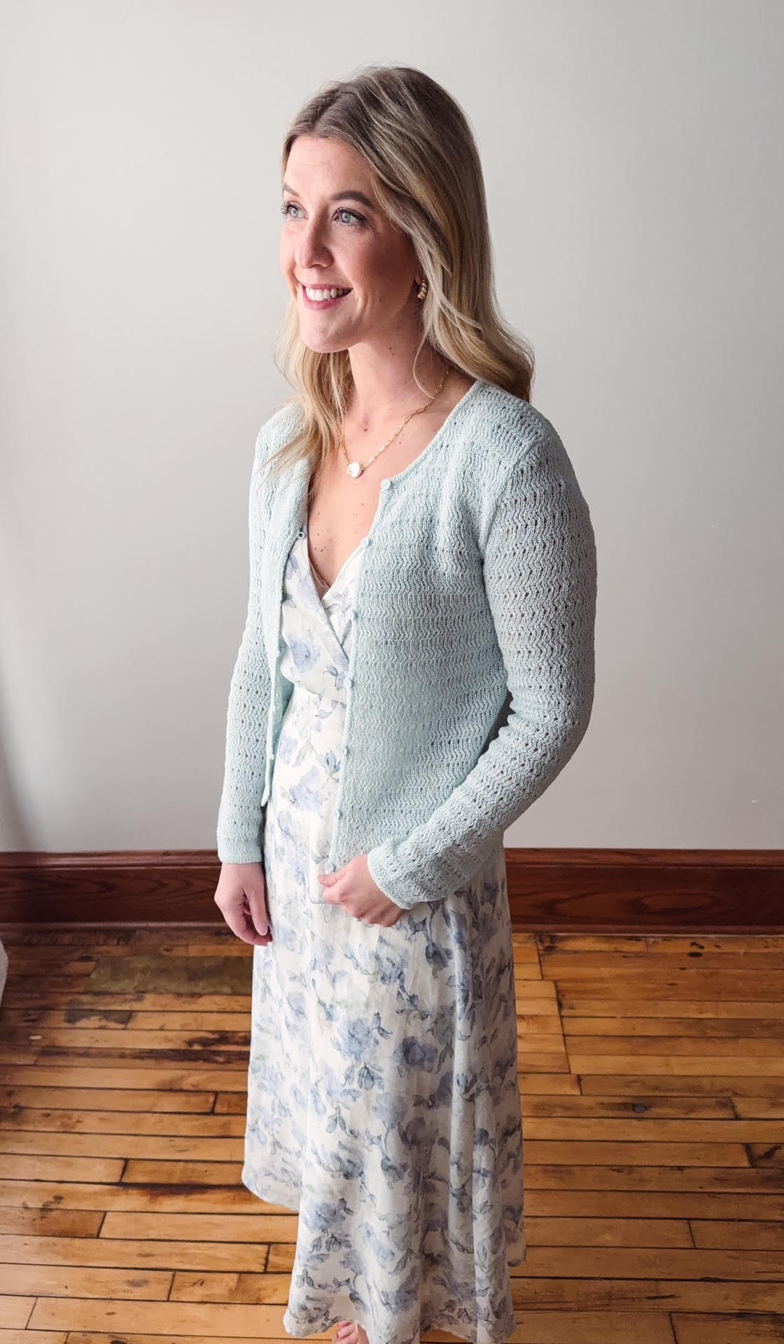 Woman wearing a light blue cardigan over a floral dress on a wooden floor.