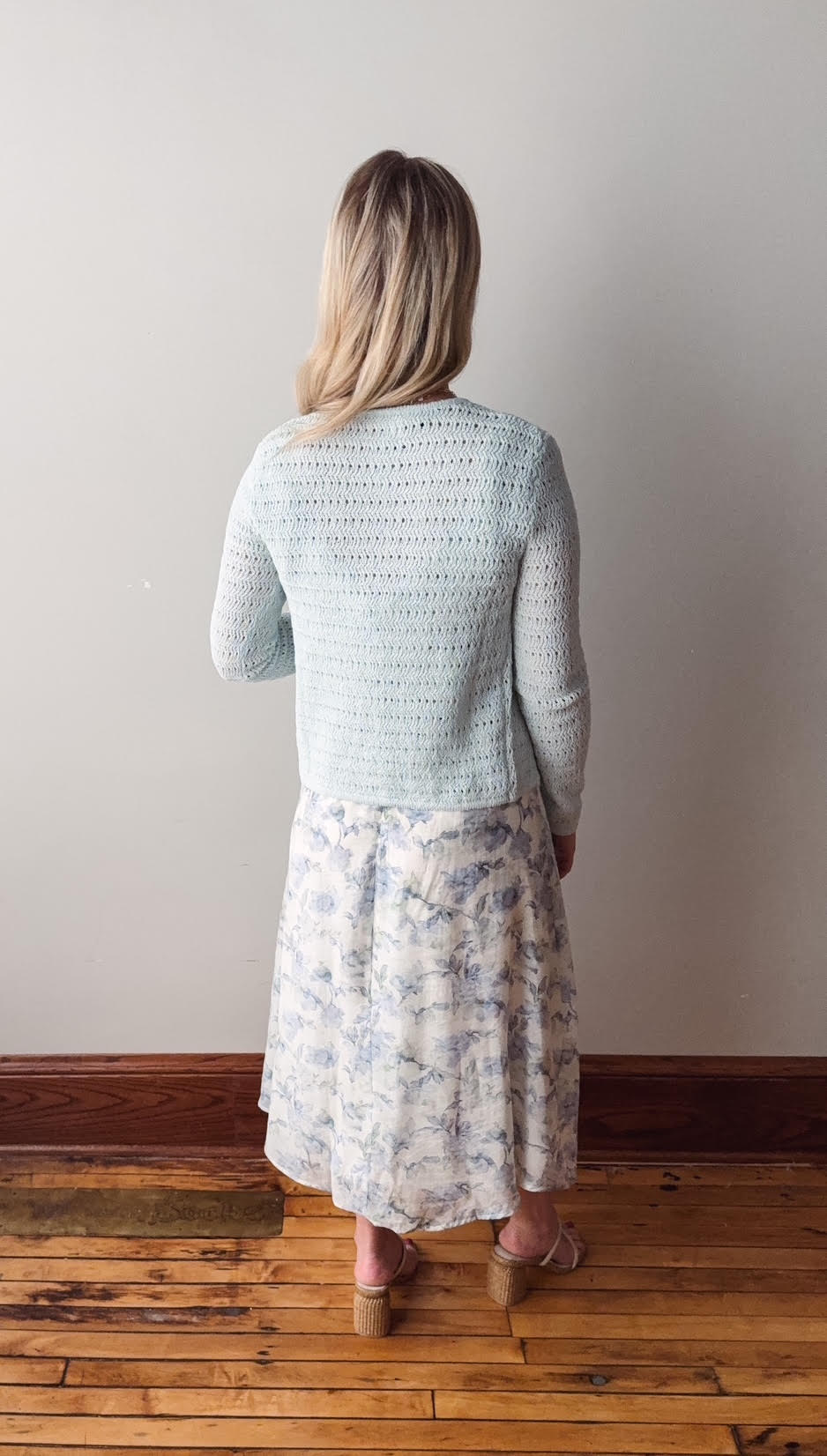 Woman wearing a light blue knitted cardigan and floral skirt standing against a plain wall.