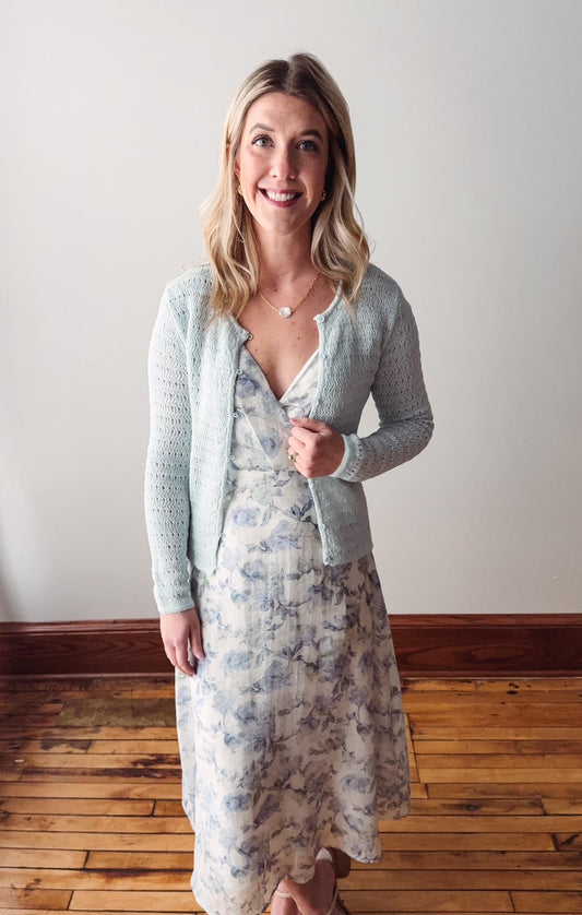 Woman wearing a light blue cardigan over a floral dress on a wooden floor.