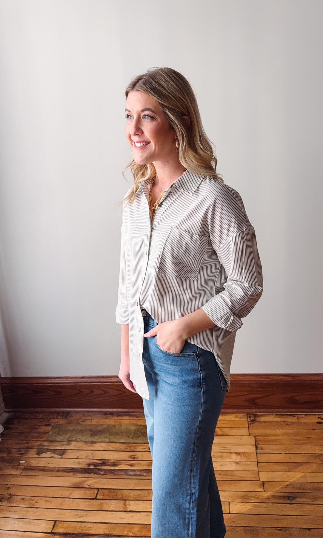 Woman wearing a light-colored button-up shirt and blue jeans standing against a white wall.