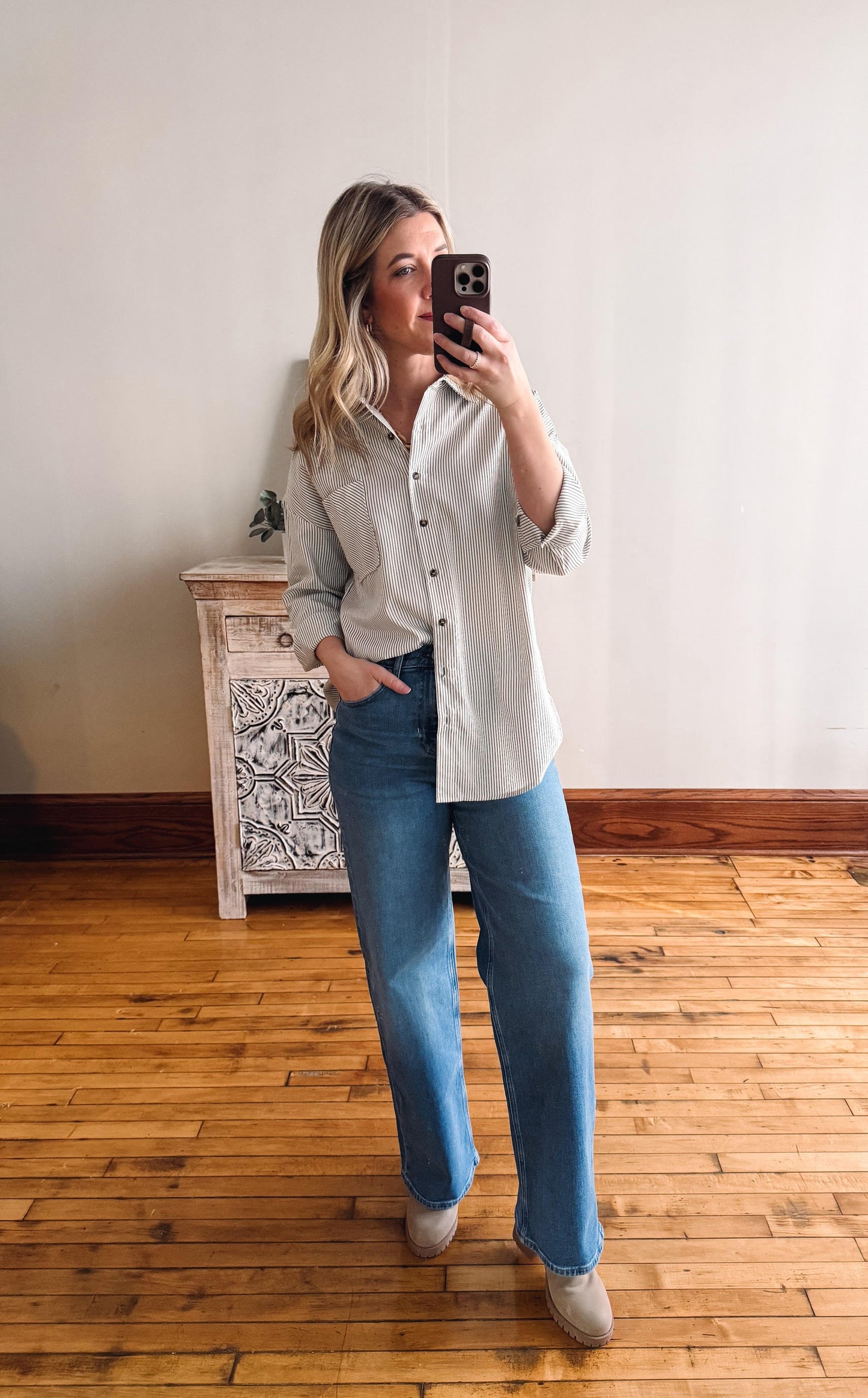 Woman taking a mirror selfie wearing a light-colored shirt and blue jeans in a room with wooden flooring.