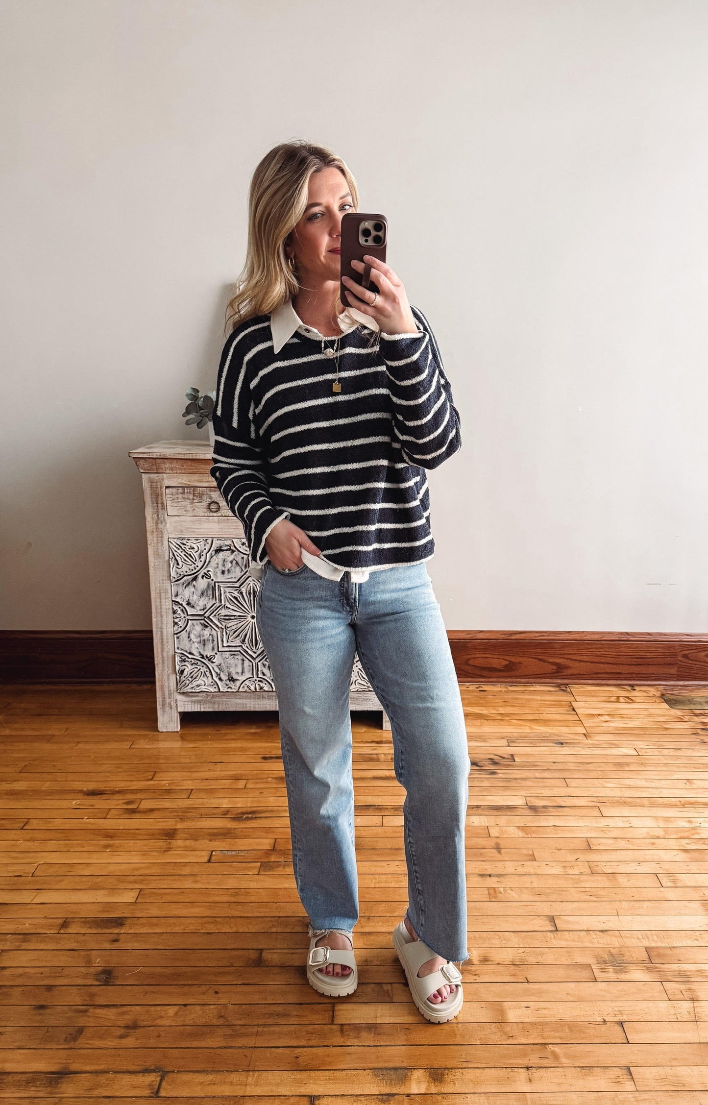 Woman taking a mirror selfie wearing a striped sweater and jeans in a room with wooden flooring.