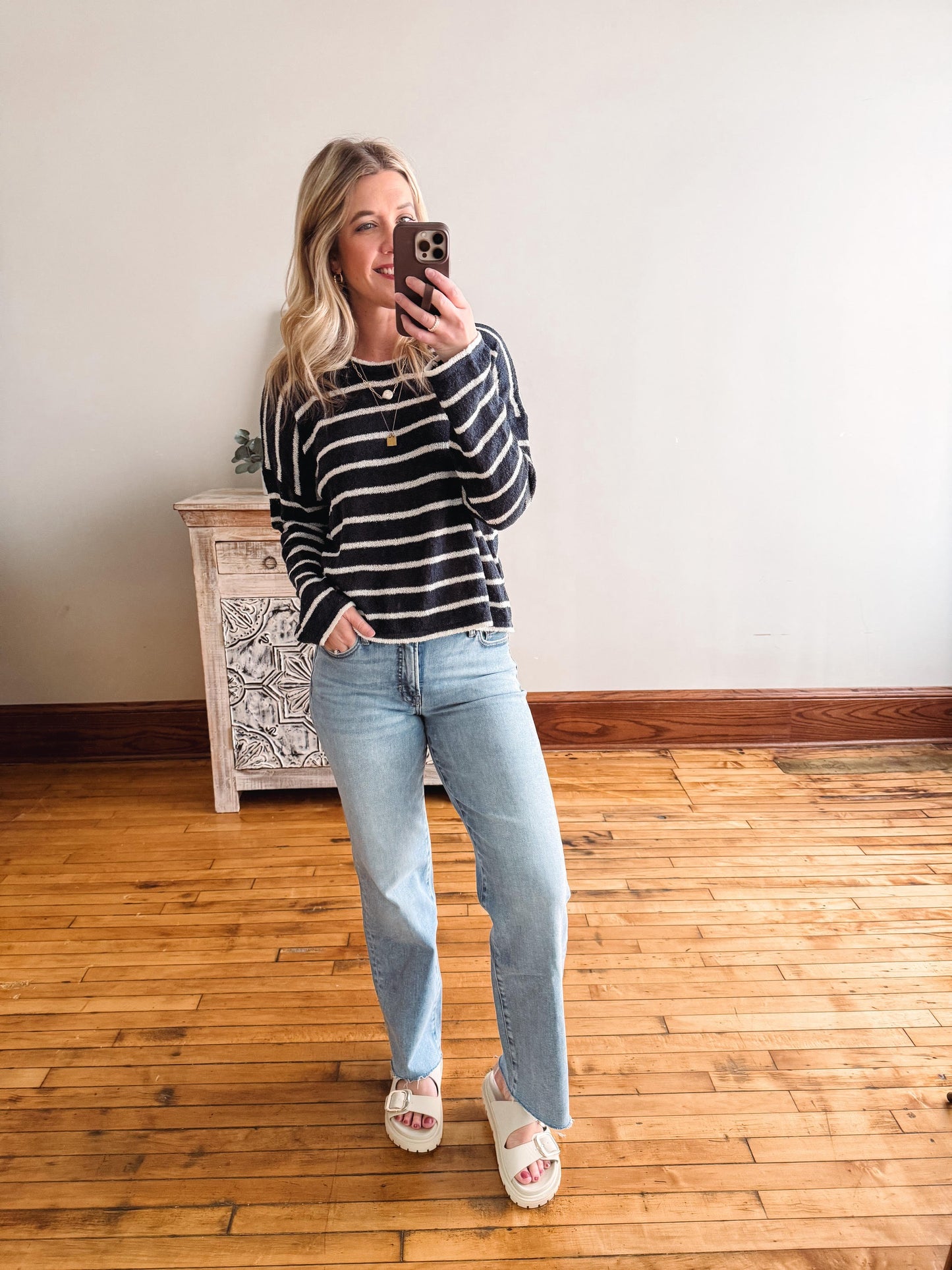 Woman taking a mirror selfie wearing a striped sweater and jeans in a room with wooden flooring.