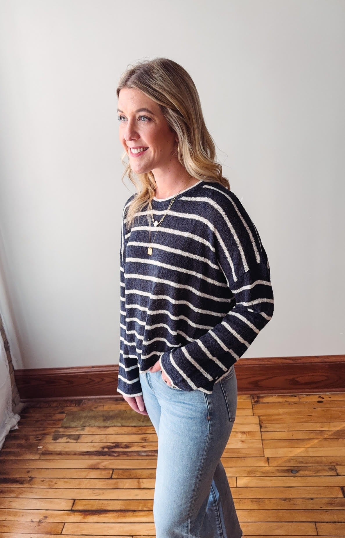 Woman wearing a black and white striped sweater and blue jeans standing in a room with wooden flooring.