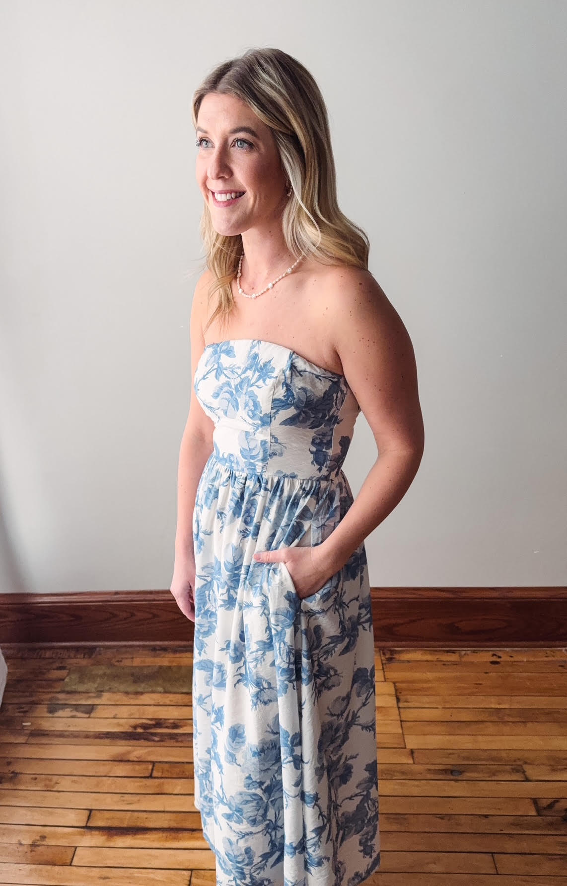 Woman wearing a strapless floral dress standing on a wooden floor with a white wall background