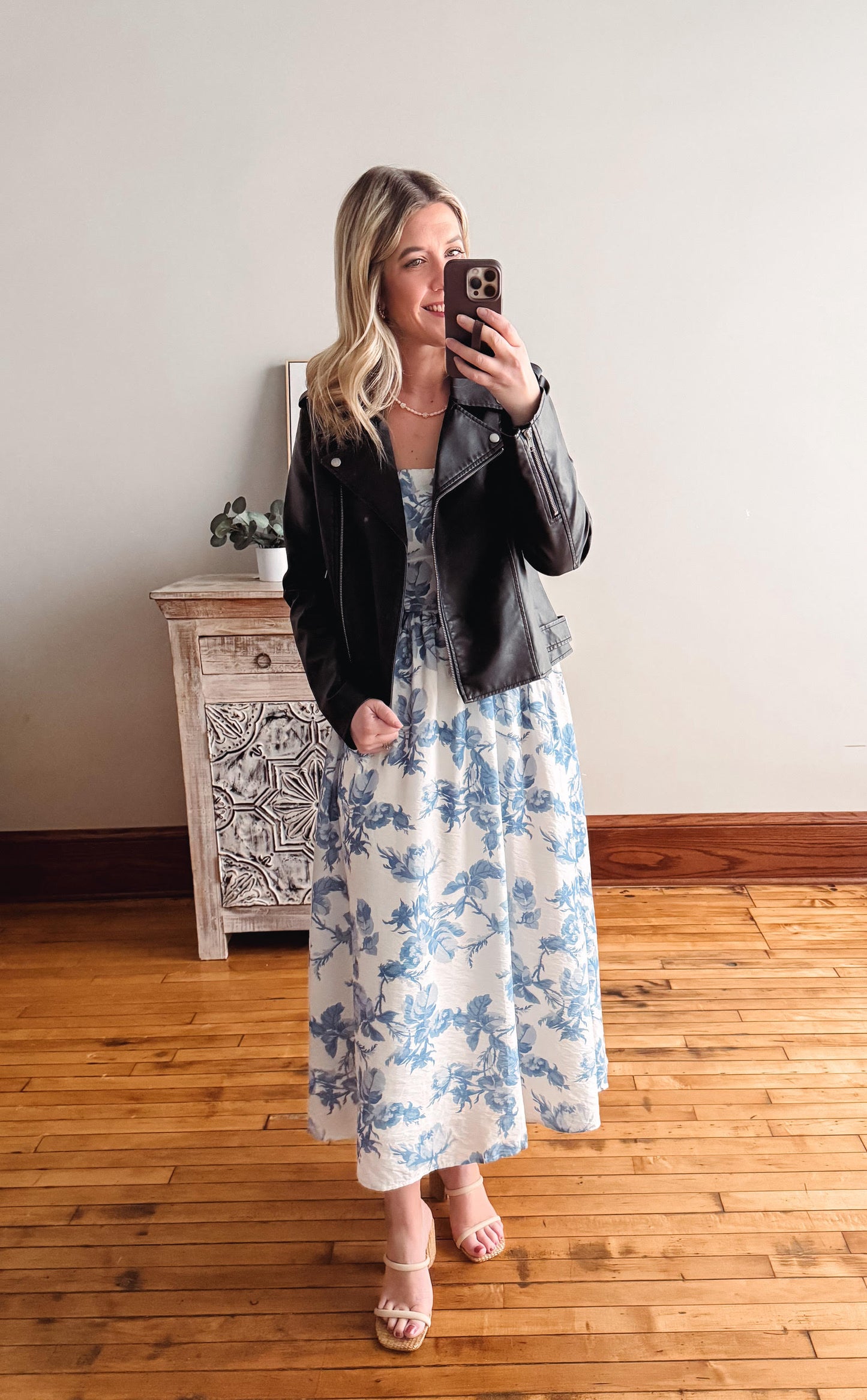 Woman taking a mirror selfie wearing a floral dress and black jacket in a room with wooden flooring.