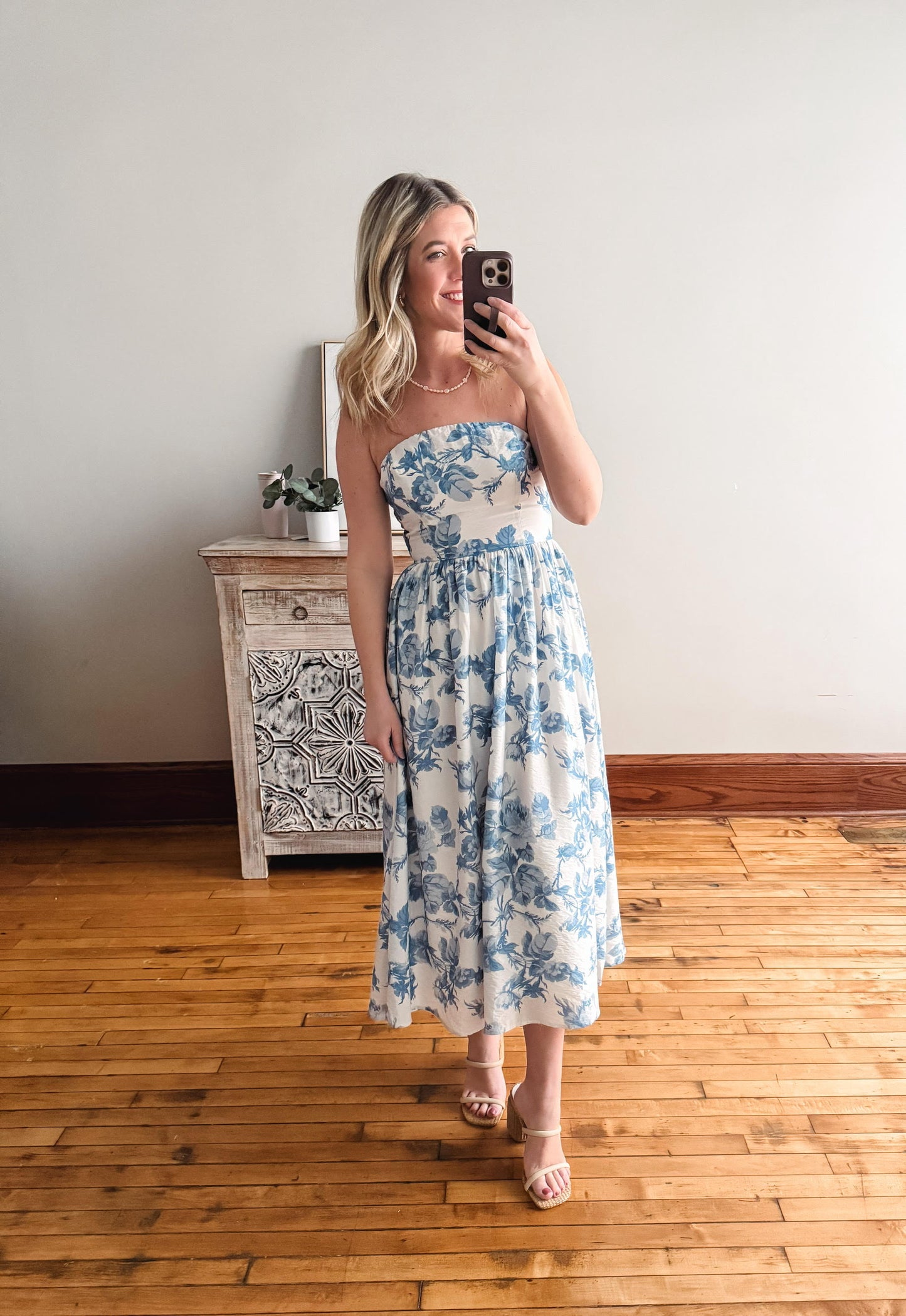 Woman taking a mirror selfie wearing a blue floral dress in a room with wooden flooring and a decorative cabinet.