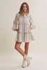 A woman modeling a long-sleeve mini dress with a beige and white floral print, blue piping details, and cream-colored ankle boots, set against a plain beige studio background.