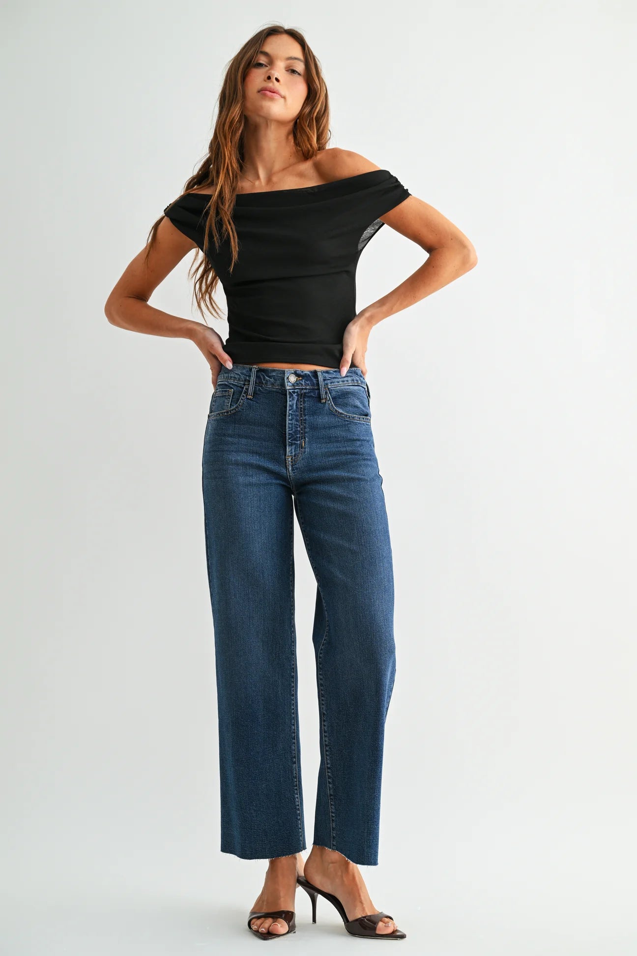 A woman modeling a black off-the-shoulder short-sleeve top and high-waisted, wide-leg blue jeans against a plain light gray studio background.