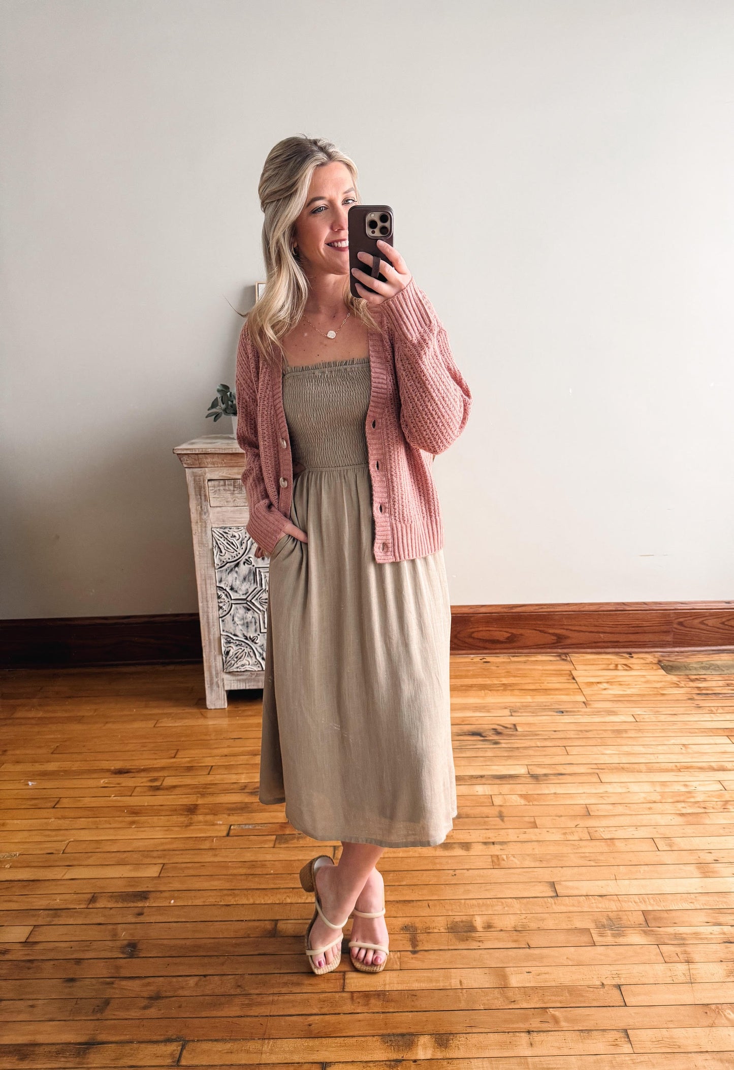 Woman taking a mirror selfie wearing a pink cardigan over a dress in a room with wooden flooring.