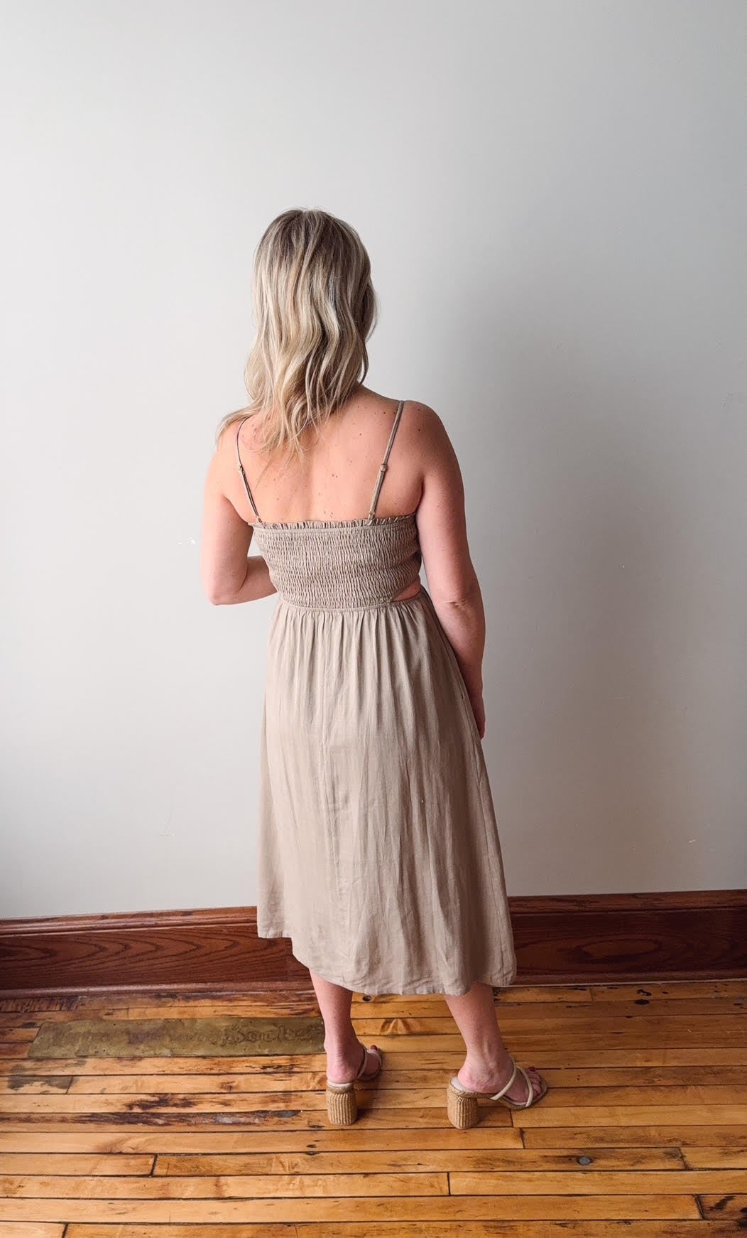 Woman wearing a beige dress standing in a room with wooden flooring and a white wall.
