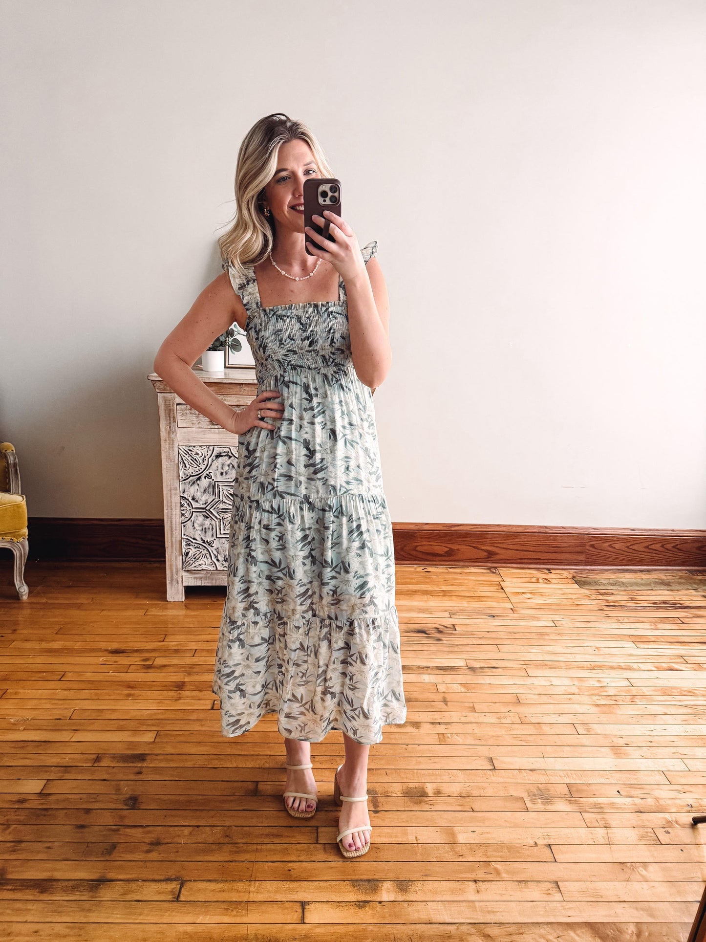 Woman taking a mirror selfie wearing a floral dress in a room with wooden flooring.